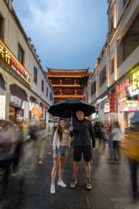 Kamy and Eric standing in an old town street under the rain, with the Guangji Gate Tower in the background.
