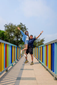 Eric lifting Kamy in the air with one arm, on a rainbow coloured bridge.