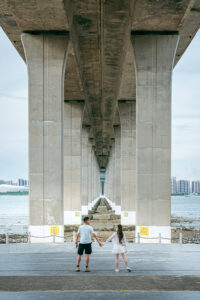 Kamy and Eric holding hands under a bridge by a bay.